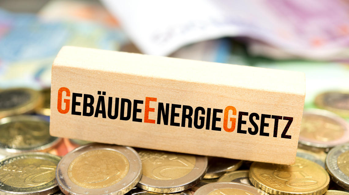 Holzblock mit der Aufschrift Gebäudeenergiegesetz auf gestapelten Euromünzen, im Hintergrund Euroscheine, als Symbol für gesetzliche Anforderungen bei der Wärmedämmung eines Gebäudes